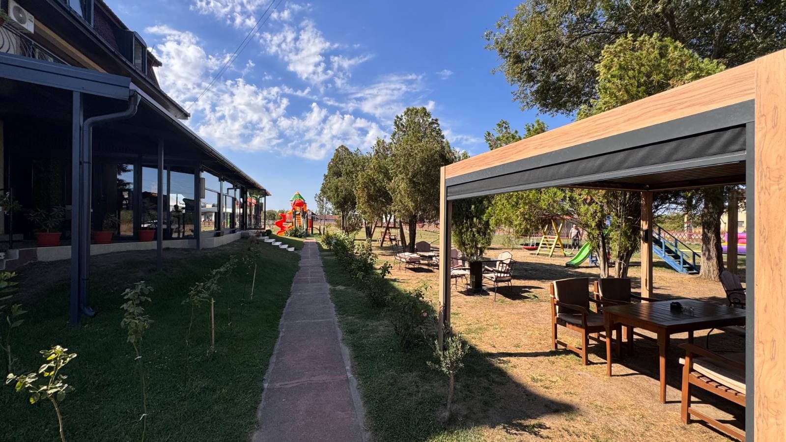 Staza između staklene bašte restorana i natkrivenog dela sa stolovima, u pozadini igralište