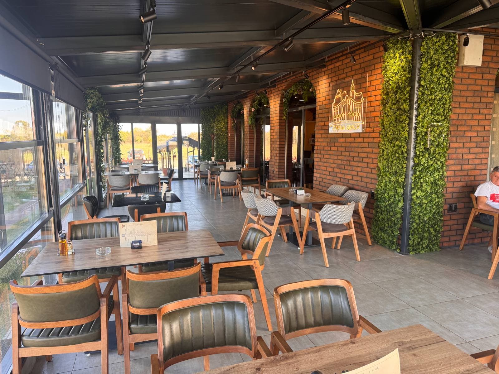 Zastakljeni deo restorana sa drvenim stolicama, zidom od opeke i zelenim zidovima